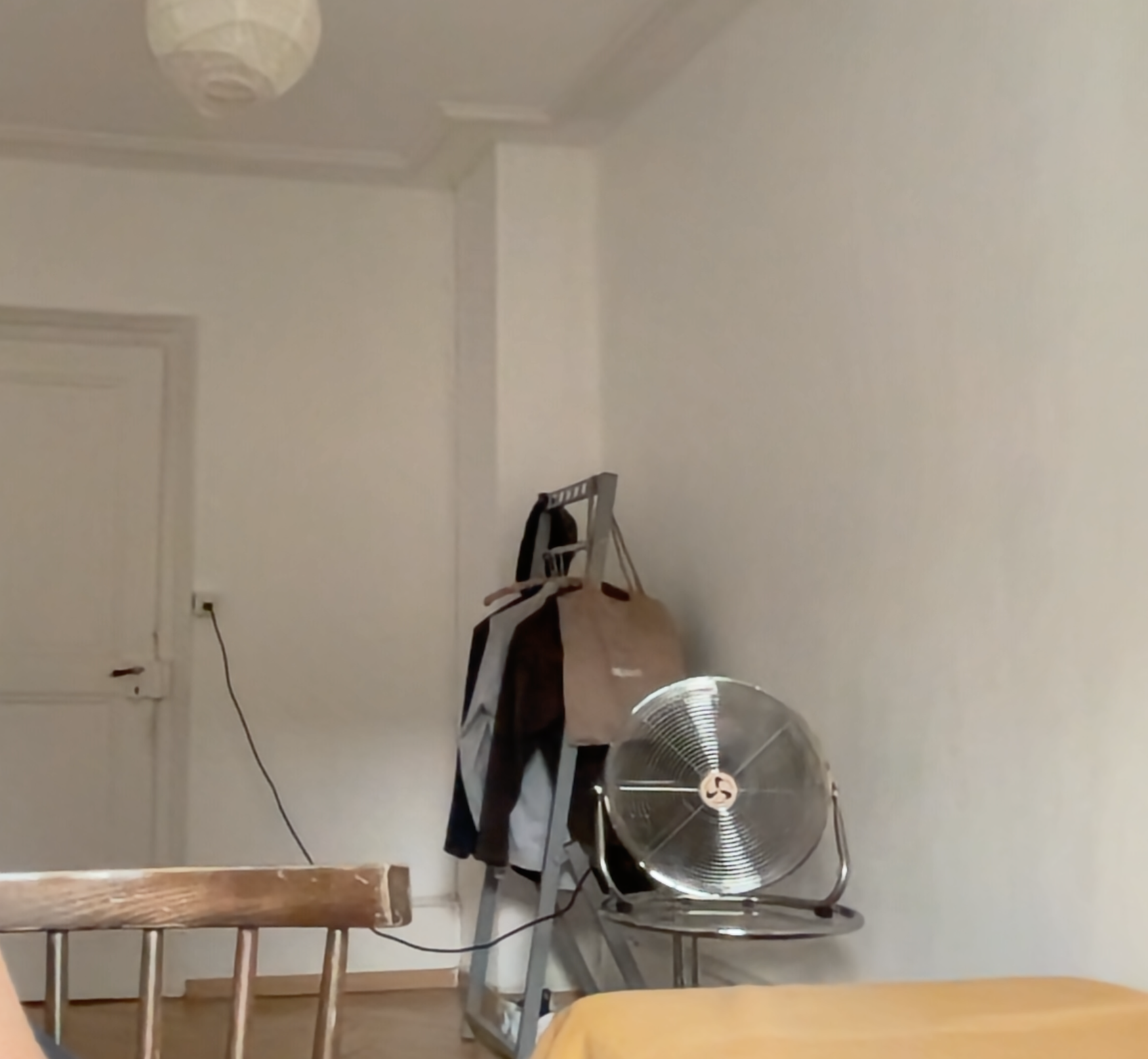 photo of a room interior with white walls, the foot of a bed with orange duvet, a fan on a small table, and a clothes rack in the back corner