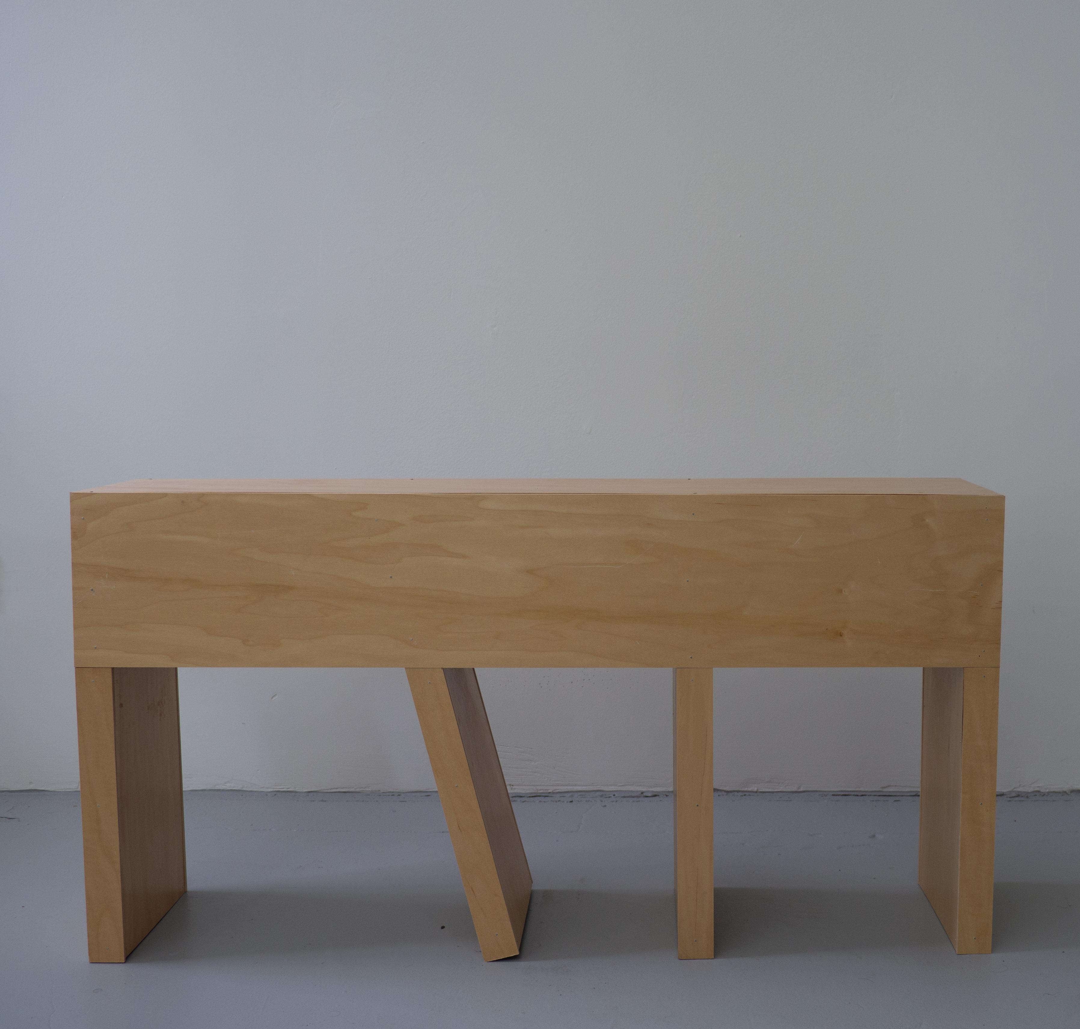 Four legged wooden bench against a white wall, sitting on a gray floor. One leg is off-kilter but still touches the ground