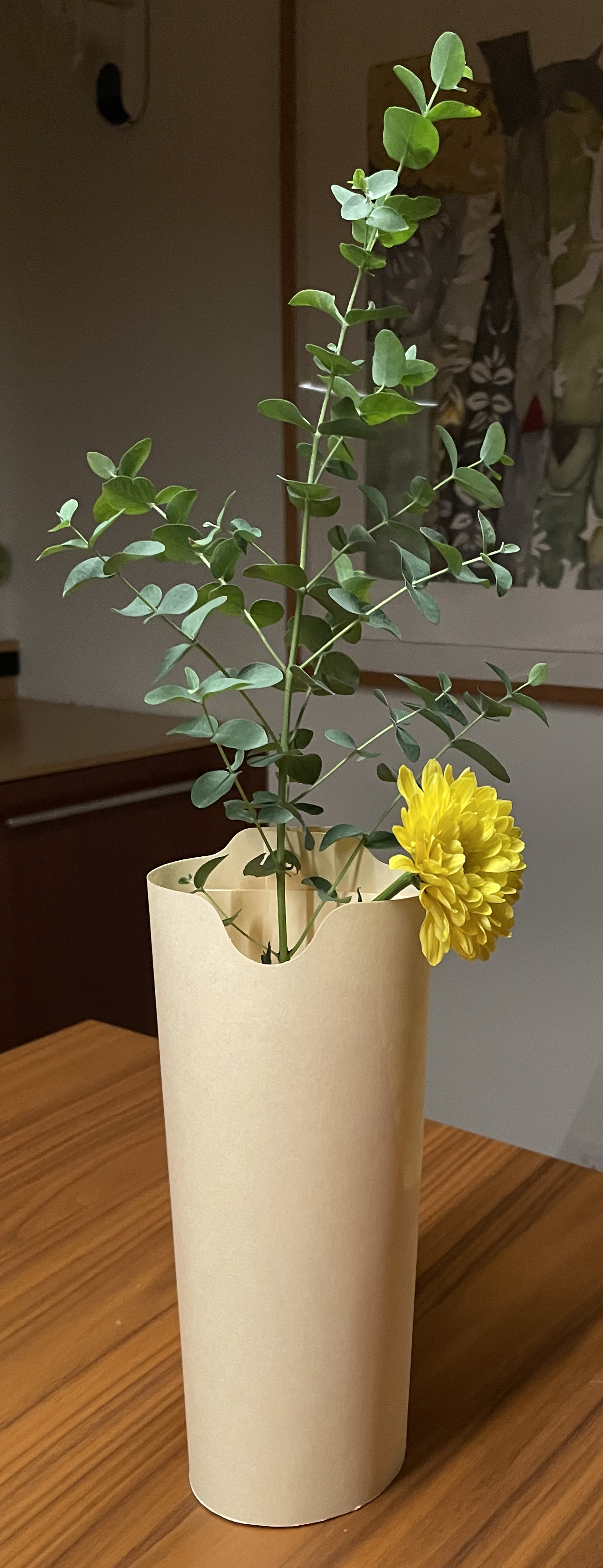 a vase made out of a manila folder sits on a kitchen counter, with a vertical frond of eucalyptus and yellow flowers emerging from the vessel