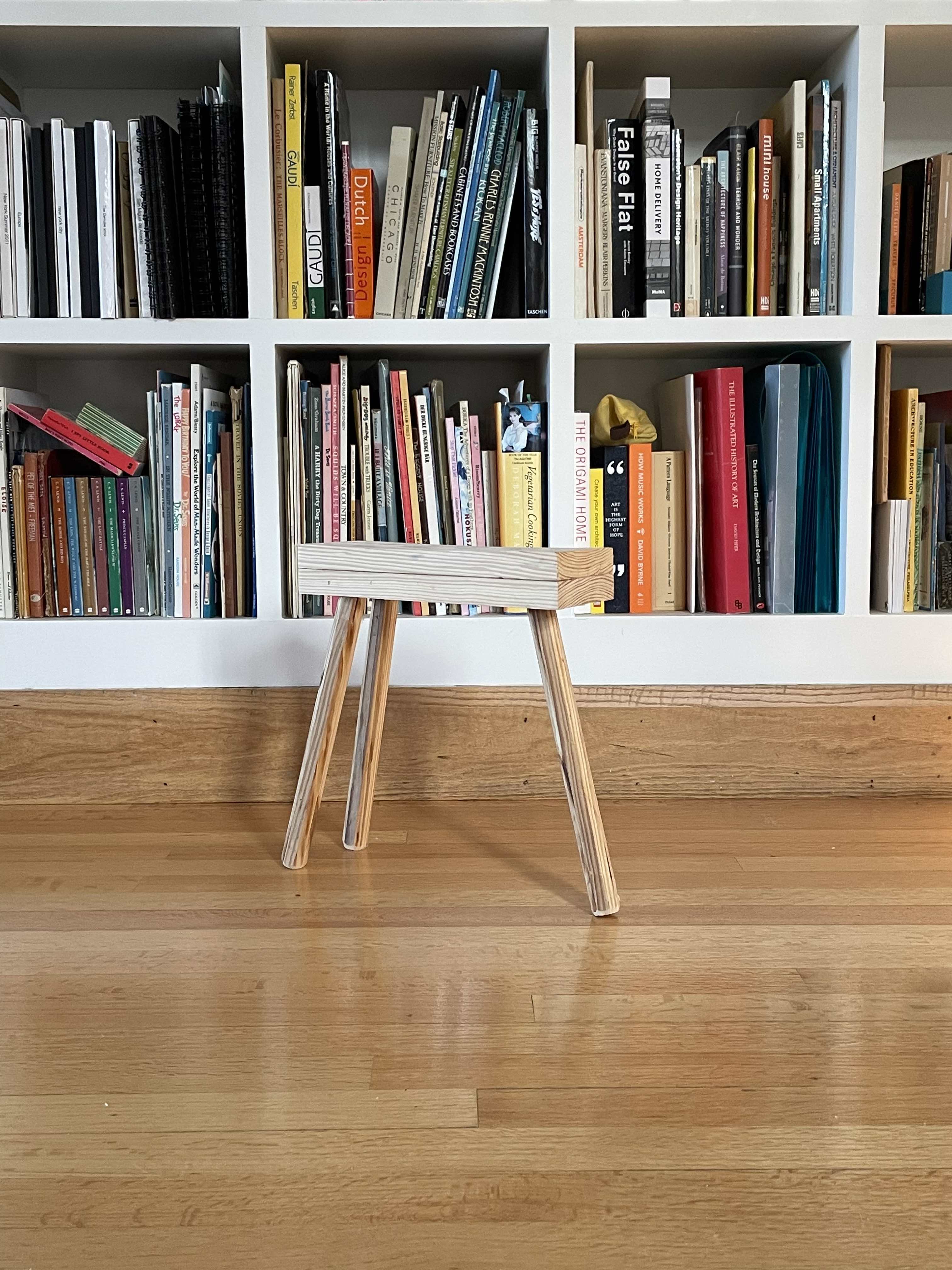 a wooden stool with three legs stands in front of a bookcase on a hardwood floor